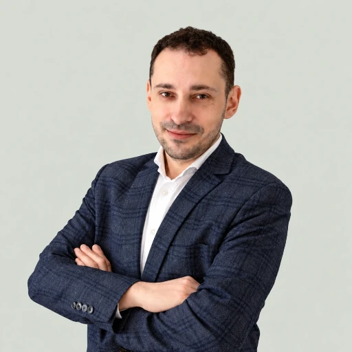 Portret Leszka Chojnickiego na jasnoszarym tle. Ma krótkie ciemne włosy i kilkudniowy zarost. Ubrany jest w granatową marynarkę w delikatną kratę i białą koszulę. Stoi z założonymi rękami, patrzy w obiektyw z lekkim uśmiechem.