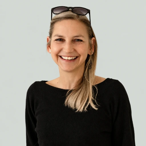 Portret Anny Karoń na jasnoszarym tle. Ma długie blond włosy przerzucone na prawe ramię, a na czubku głowy okulary przeciwsłoneczne. Ubrana jest w czarny sweter z szerokim dekoltem. Szeroko się uśmiecha, patrząc w obiektyw.